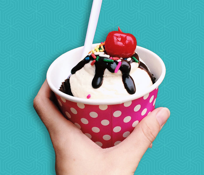 Hand holding a cup of ice cream, which is topped with chocolate syrup, sprinkles, and a cherry