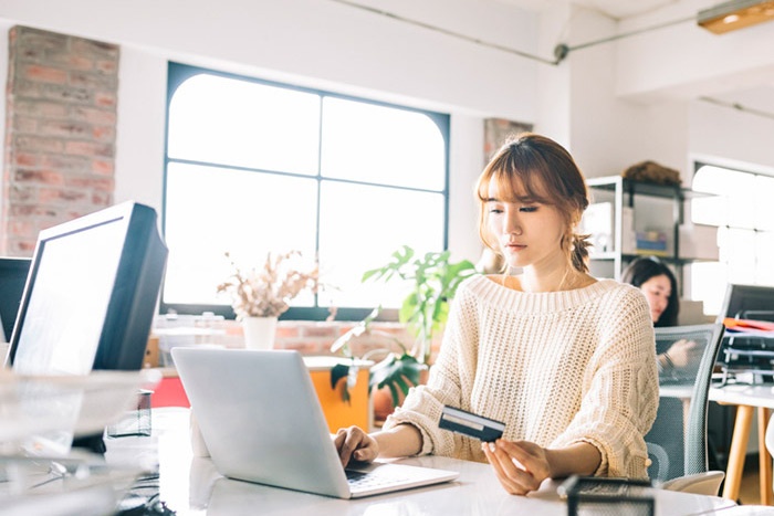 Young woman sitting at office desk while looking at laptop and holding debit card