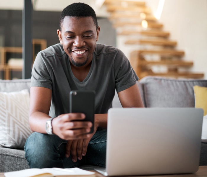 man smiling at phone while sitting on couch in front of table with laptop and papers