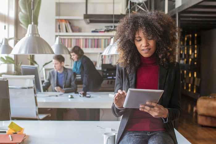 Woman in office setting sitting on side of desk while typing on tablet