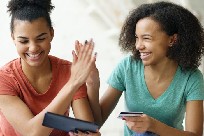 Young adult women high fiving while holding a bank card while looking at a tablet